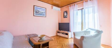 Uma sala de estar aconchegante com um sofá, um rack para televisão, uma mesa de jantar com uma tigela e uma poltrona de cor clara.