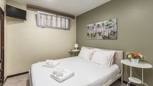 Habitación de hotel con cama, televisor montado en la pared, ventana con persianas, un cuadro, una mesita con lámpara y un arreglo floral.