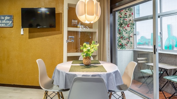 Un área de comedor con una mesa redonda, cuatro sillas, un televisor en la pared y un amplio ventanal con vistas a la ciudad.