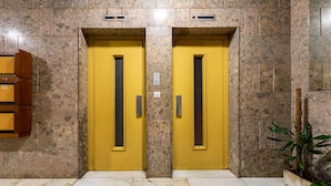 Dos puertas de ascensor amarillas con un panel texturizado, ubicadas en un vestíbulo con paredes de piedra y piso de baldosas.