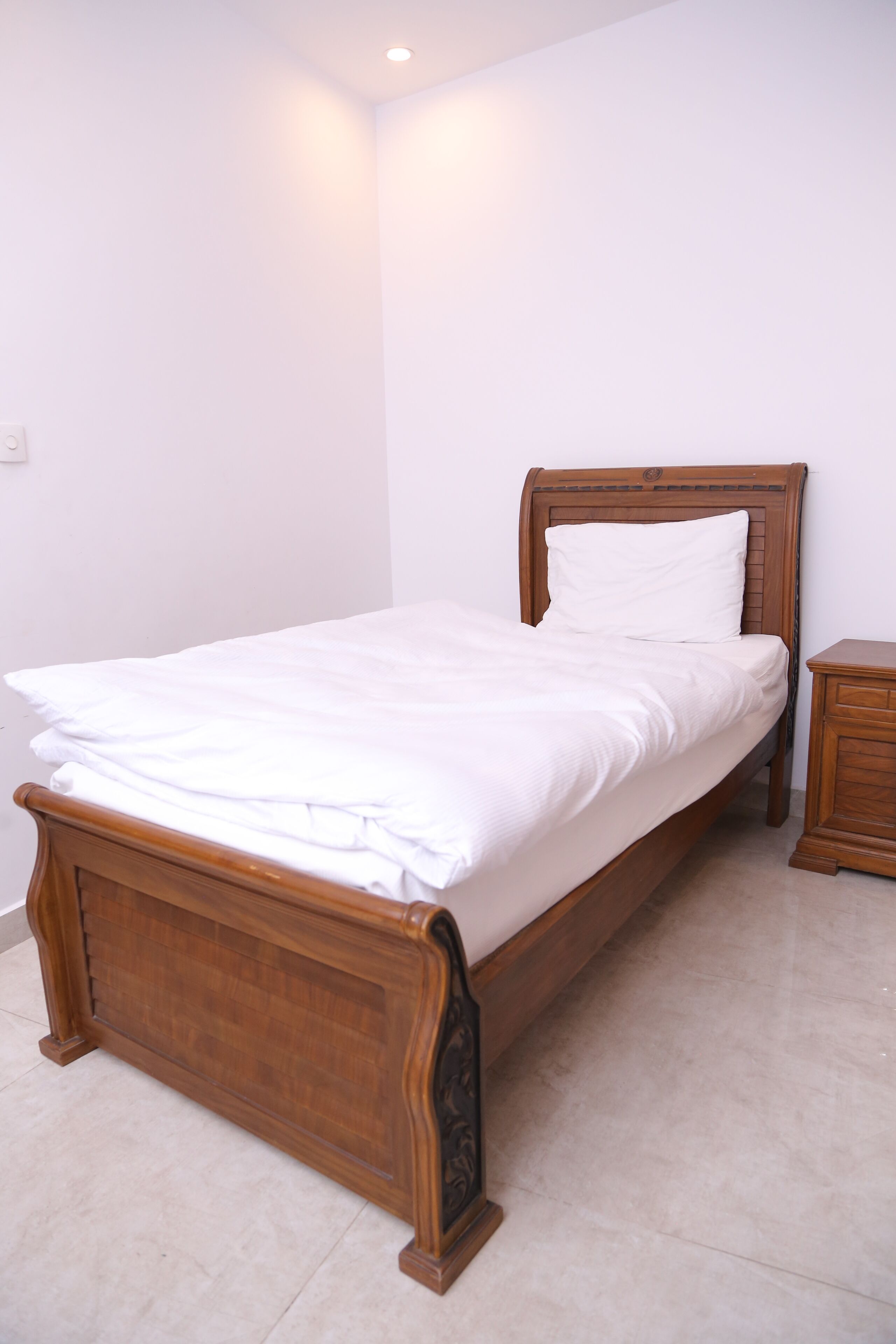 A neatly made bed with a wooden frame and white bedding in a room with white walls and a tiled floor.