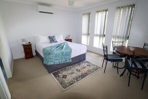 Un dormitorio con cama, mesitas de noche, una mesa de comedor con sillas y una ventana con persianas.