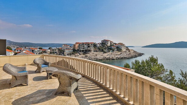 A balcony with wicker furniture overlooking a coastal town with buildings and a body of water.