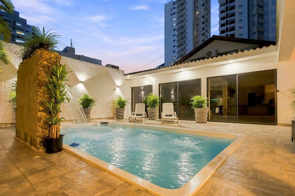 An outdoor swimming pool with a stone wall, potted plants, and lounge chairs.