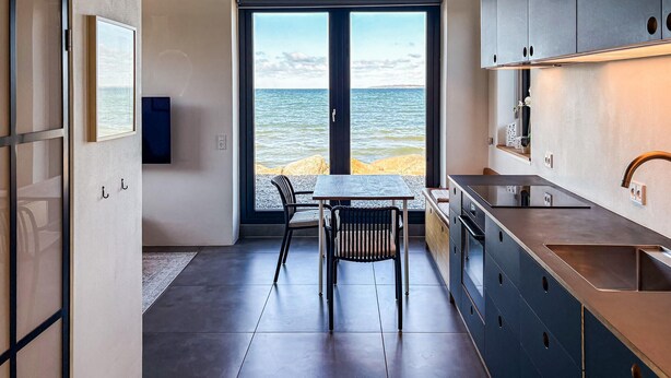 Ett modernt kök med utsikt över havet, en matplats med stolar och ett stort fönster.