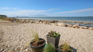 En sandstrand med klippformationer och fri utsikt över havet. I förgrunden finns planterade blommor i krukor.
