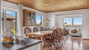 A dining area with a wooden table and chairs, a cabinet with glass doors, and a view of the outdoors through large windows. - Gorgeous home in Læsø with kitchen (Læsø)