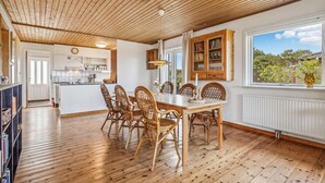A dining area with a wooden table and chairs, a kitchen in the background, and a window with a view of greenery. - Gorgeous home in Læsø with kitchen (Læsø)