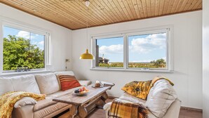 A cozy room with a wooden ceiling, two large windows offering a view of greenery and a distant building, a wooden table with a fruit bowl, and a sofa with a patterned throw blanket. - Gorgeous home in Læsø with kitchen (Læsø)