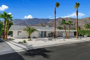 A modern two-story building with a flat roof, surrounded by palm trees and cacti, with mountains in the background.