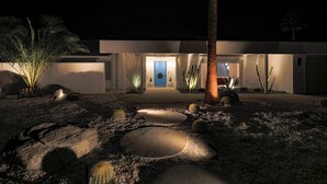 Una casa moderna con una entrada bien iluminada, un camino que lleva a la puerta y un jardín desértico con rocas y cactus.