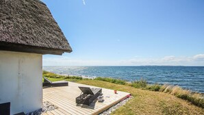 A wooden deck with a thatched-roof building, overlooking a calm sea. - Gorgeous home in Samsø with sauna (Samsø)