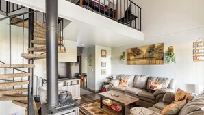Un salón amplio con un sofá grande, una mesita de centro y una chimenea. Se observa una escalera de caracol al fondo.
