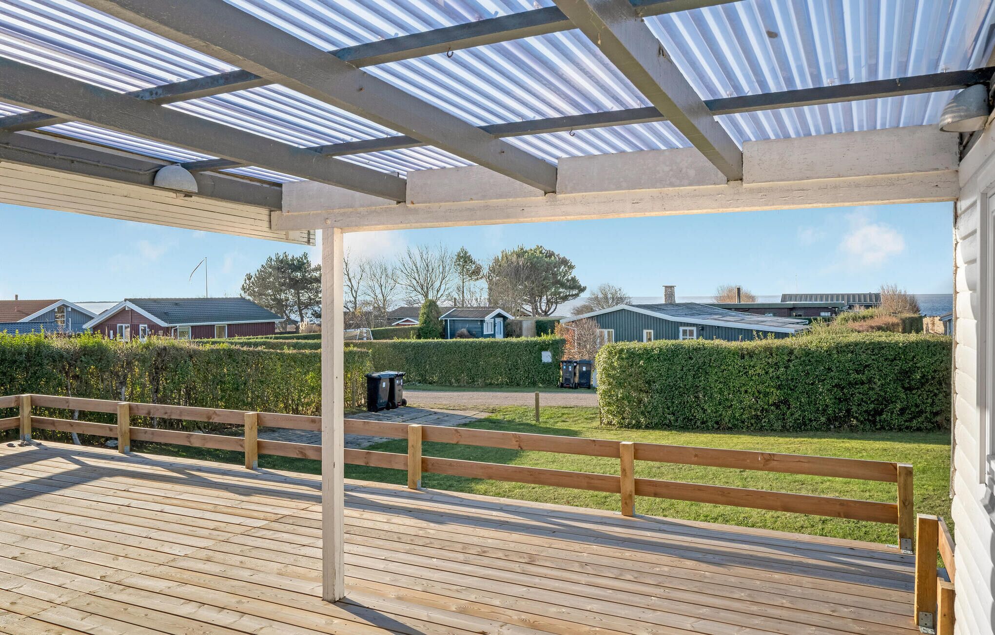 Een overdekt buitengebied met een houten terras, een metalen dak en uitzicht op woongebouwen en een waterpartij.