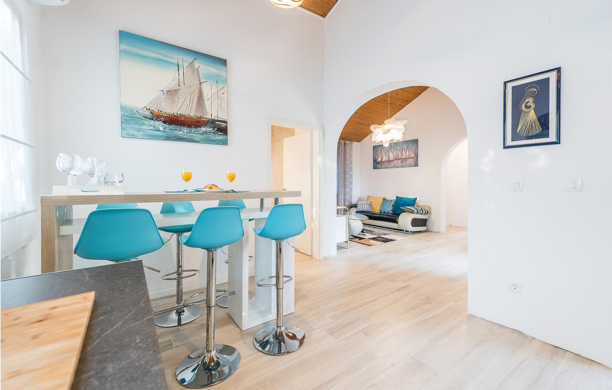 A modern kitchen with a dining area, featuring a wooden table, blue chairs, and a painting of a sailboat.
