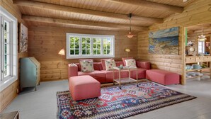 Ein gemütliches Blockhaus-Interieur mit einer rosa Sofagarnitur, einem gemusterten Teppich und einem großen Fenster mit Blick auf Grünflächen.