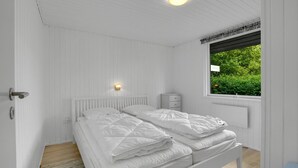 A bedroom with a bed, a bedside table, a nightstand, a window with a view of greenery, and a ceiling light.