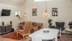 Een moderne woonkamer met een hoge plafond, een flatscreen-tv, een eettafel en twee stoelen. Er zijn twee hangende pendellampen en een potplant.