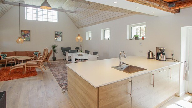 Een ruime keuken met een groot eiland, houten keukenkastjes en een rustiek plafond. Er is een eethoek met een tafel en stoelen, en een zithoek met een bank en een vloerkleed.