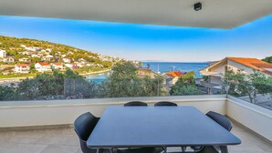Un balcón con mesa y sillas, con vista a un pueblo costero con casas y un cielo azul despejado.