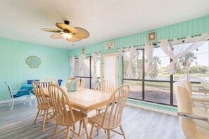 Un comedor con una mesa y sillas de madera, un ventilador de techo y vistas a palmeras a través de amplios ventanales.
