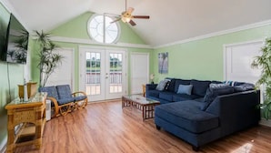 A spacious living room with a high ceiling, a ceiling fan, a large window, a dark blue sofa, a wicker chair, and a small coffee table. - Cheerful 2 Bedroom Cottage with fishing deck! (Bayview)
