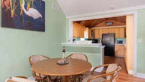 A dining area with a round wooden table and wicker chairs, a kitchen with wooden cabinets and a black refrigerator, and a painting of a white bird on the wall. - Cheerful 2 Bedroom Cottage with fishing deck! (Bayview)