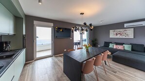 Una cocina moderna con una puerta corrediza que da a un balcón, una zona de comedor con una mesa y sillas oscuras, y una sala de estar con un sofá y un cuadro en la pared.
