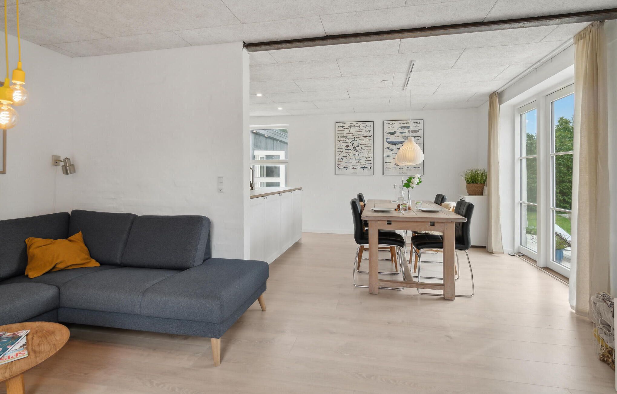 Un salon moderne avec un canapé gris, une table basse en bois et un coin repas comprenant une table et des chaises en bois. On y trouve deux appliques murales et une grande fenêtre avec des rideaux.
