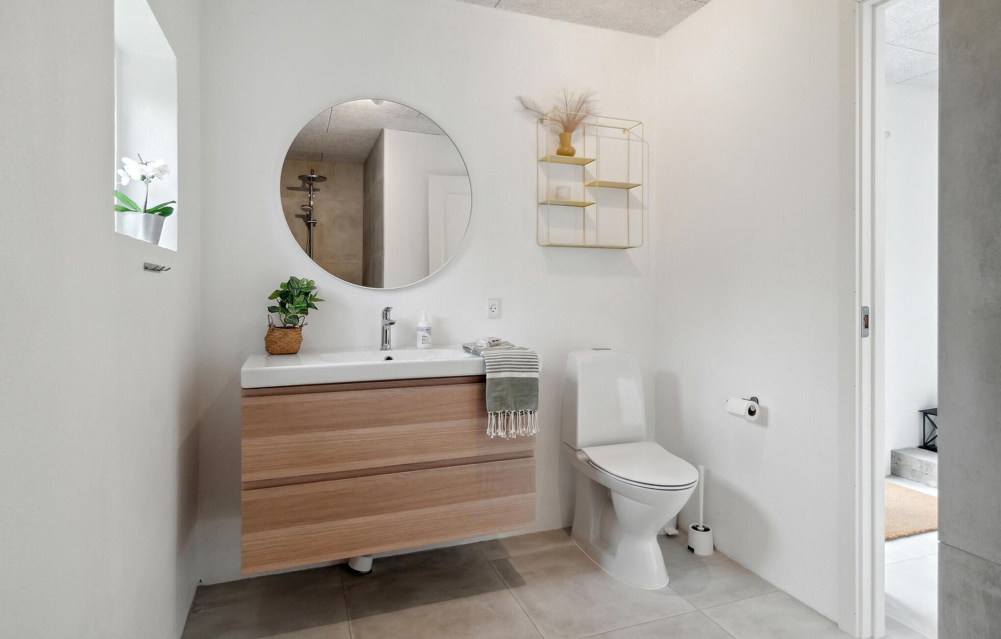 A modern bathroom with a wooden vanity, a round mirror, a toilet, and a small plant on the vanity.
