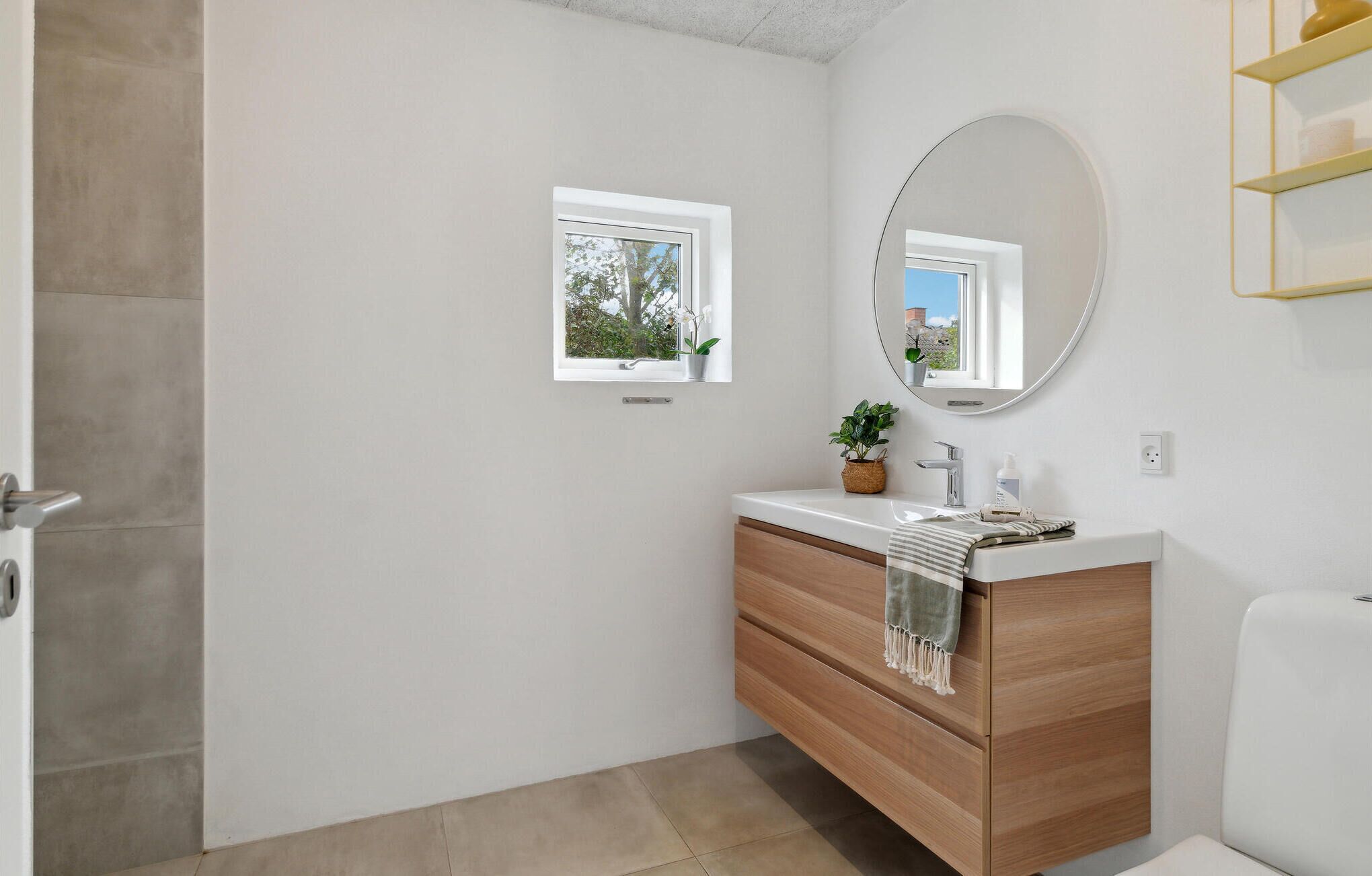 Une salle de bain moderne avec un meuble-lavabo en bois, un miroir rond, une petite fenêtre et une plante en pot.