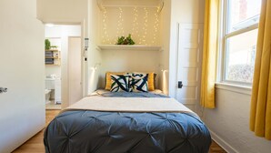 A bedroom with a bed, a window with yellow curtains, a shelf with plants, and a wall-mounted light fixture. - Charming 1BR Basecamp | Close to UofA (Tucson)