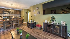 Eine moderne Küche mit Holzausstattung, ein Essbereich mit Holztisch und -stühlen sowie ein Wohnzimmer mit dunklem Holzboden, einem Couchtisch und einem kleinen Weihnachtsbaum.
