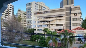 Un edificio residencial de varios pisos con balcones, cielo despejado y algo de vegetación en primer plano.