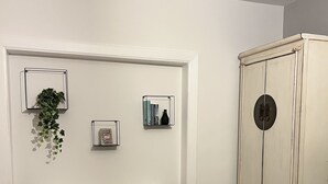 Une pièce avec un mur blanc, un petit bureau blanc orné d’une plante, un pouf noir et une armoire blanche.