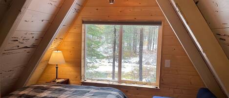 A cozy attic bedroom with a large window, a bed, a blue chair, and a hanging light fixture.