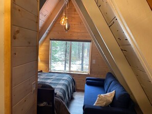Um quarto de sótão acolhedor, com uma cama, um sofá azul e uma janela grande com vista para a floresta.