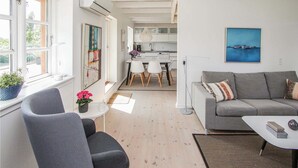 A modern living room with a grey sofa, a small table, and a chair. There is a kitchen in the background with white cabinets and a dining area with a white table and chairs. - Cozy apartment in Samsø with kitchen (Samsø)