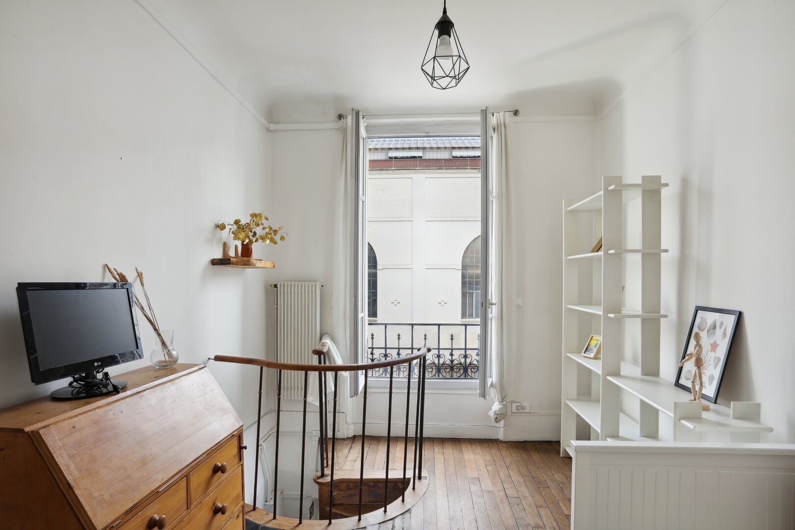 A room with a wooden desk, a black monitor, a radiator, a white radiator, a wooden staircase, a white bookshelf, and a window with a view of buildings.