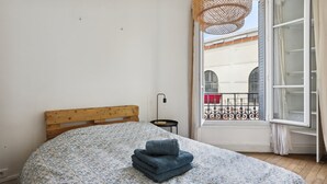 Quarto com cabeceira de madeira, colcha floral, pilha de toalhas, abajur e vista para uma varanda com grade e um prédio ao fundo.