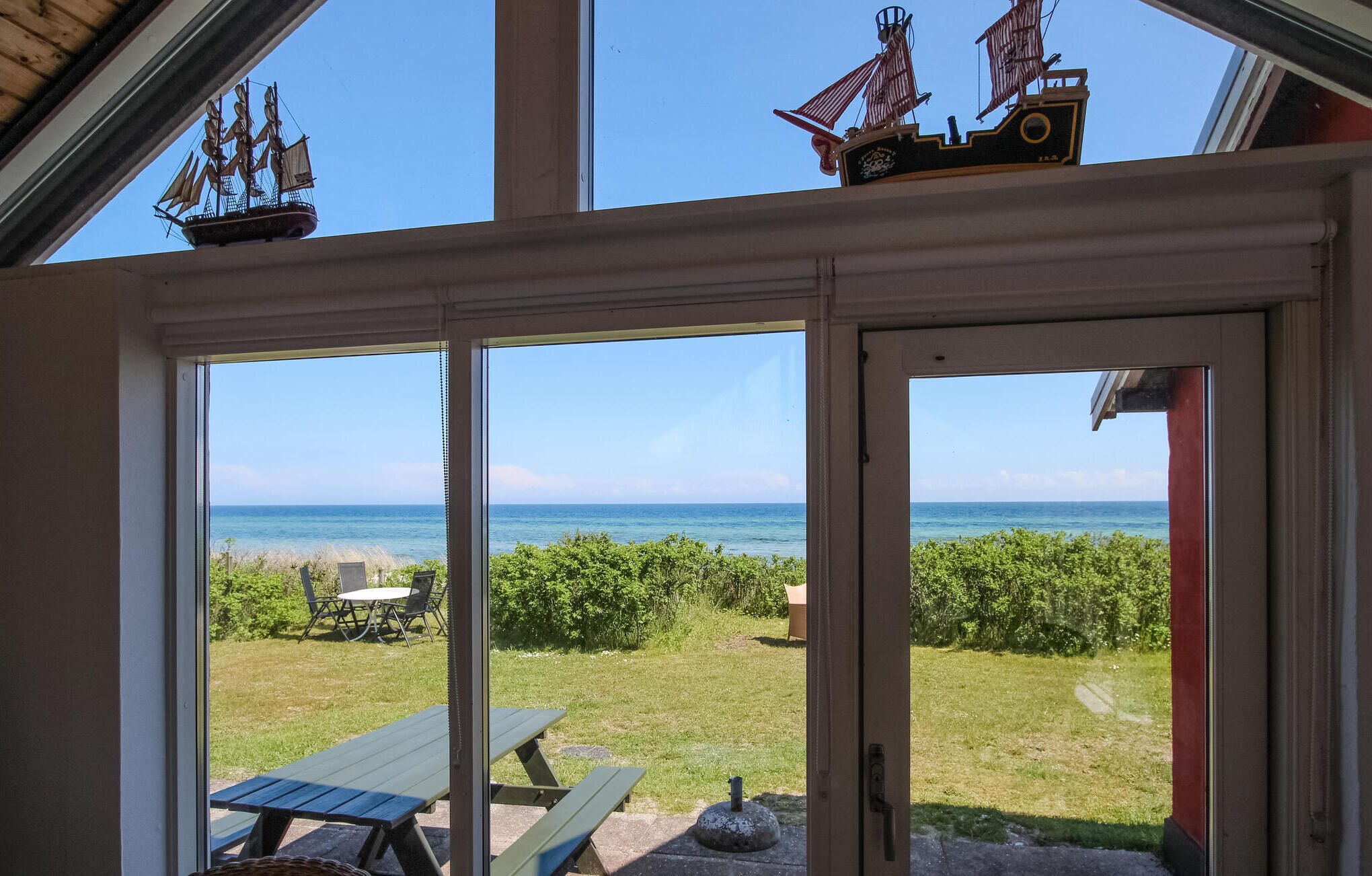 Ein Zimmer mit großen Fenstern, das einen Blick auf eine Wiese und das Meer bietet, mit Picknicktisch und Schiffsmodell.