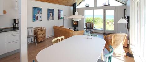 A modern kitchen with white cabinets, a dining table, wicker furniture, and a large window overlooking the sea.