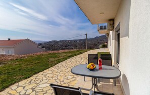 Un balcon avec une table dressée, ornée de fruits et d’une bouteille, donnant sur une place pavée de pierres et une colline.