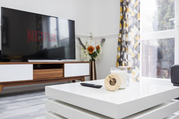 A modern living room with a flat-screen TV displaying 'NETFLIX', a remote control, and a candle on a white coffee table.