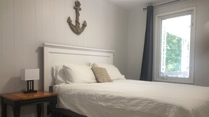 A bedroom with a bed, a nightstand, a lamp, a window with curtains, and a wall-mounted anchor decoration.
