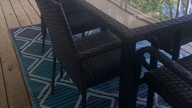 A covered outdoor seating area with a glass table and wicker chairs, surrounded by a wooden deck and a patterned rug.