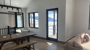 A modern interior with a wooden dining table, bench, and a sofa. There are large windows offering a view of mountains.