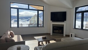 A living room with a fireplace, large windows with a view of a house and mountains, a flat-screen TV mounted above the fireplace, and a sofa.