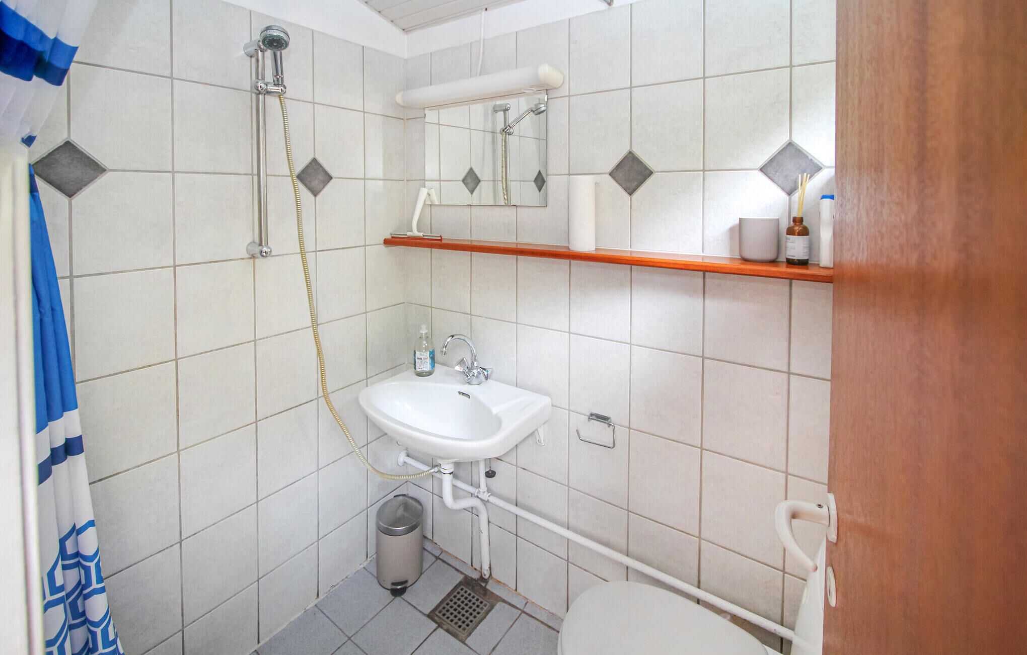 A small bathroom with a white sink, a shower, and a toilet. There is a mirror above the sink and a shelf with toiletries.