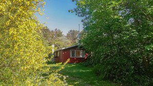 Punainen mökki, jossa on ikkuna ja savupiippu, ja sen ympärillä on vihreää ruohoa ja puita.